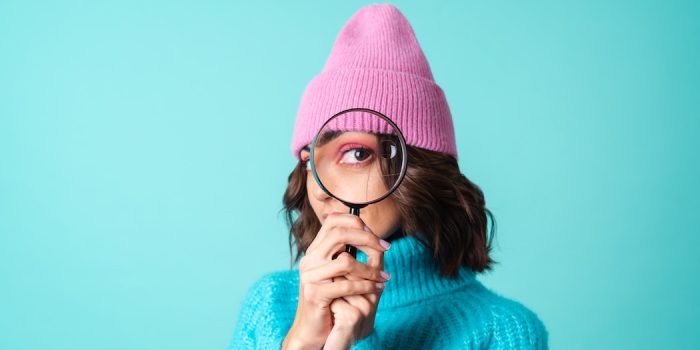 Cozy portrait of a young woman in a knitted blue sweater and a pink hat with bright makeup holding a magnifying glass, fooling around, having fun