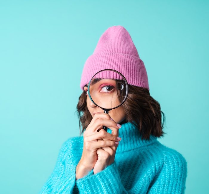 Cozy portrait of a young woman in a knitted blue sweater and a pink hat with bright makeup holding a magnifying glass, fooling around, having fun